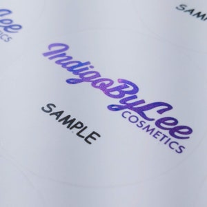 May include: A white sticker with the text "Indigo By Lee Cosmetics" in a purple and blue gradient font. The text "SAMPLE" is printed in black below the logo.