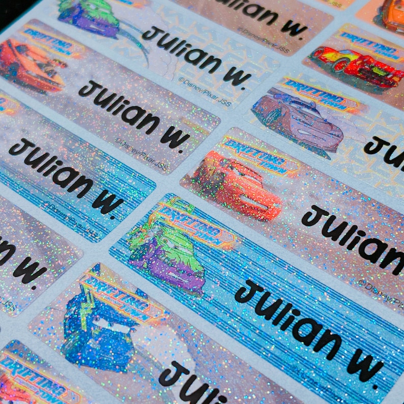 May include: A close-up of a sheet of colorful, glittery name labels with the name "Julian W." printed on each label. The labels feature a cartoon car design with a blue and red color scheme.