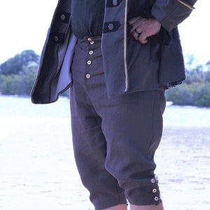 May include: A dark gray pirate-style outfit. The ensemble includes knee-length breeches with button details and a matching jacket with gold trim and black buttons. The outfit is suitable for historical reenactments or themed events.