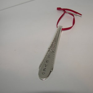 May include: A silver spoon ornament with the word "LOVE" engraved on it. The spoon has a red ribbon attached to the top for hanging.