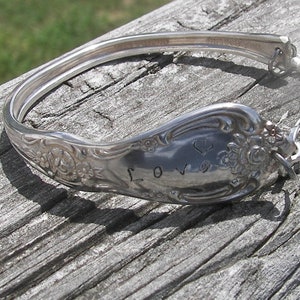 May include: A silver spoon bracelet with an ornate design and the word "LOVE" engraved on it. The bracelet is made from a repurposed spoon and has a chain closure.