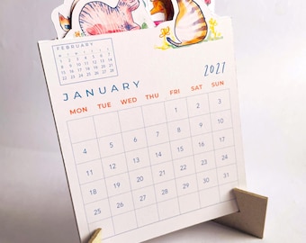 Könnte beinhalten: Ein Tischkalender für 2027 mit einem Januar-Kalenderblatt und einer kleinen Februar-Übersicht. Der obere Rand ist mit bunten Aquarellillustrationen von Katzen versehen. Der Kalender steht auf zwei hellbraunen Holzständern.