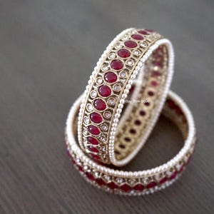 May include: Two gold-toned bangles with a repeating pattern of red gemstones and small white beads. The bangles are stacked on top of each other, with one partially inside the other. The background is a dark gray surface.