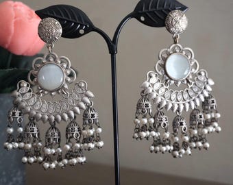 May include: Silver-toned chandelier earrings with a white stone center and dangling pearl accents. The earrings have a floral design around the central stone and small bell-shaped charms. The earrings are displayed on a black stand.