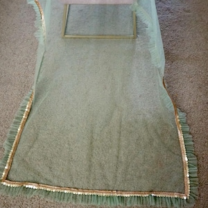 May include: A light green sheer fabric with a gold and white beaded trim. The fabric is draped over a stool.