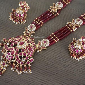 May include: A three-piece jewelry set featuring a necklace, earrings, and a ring. The necklace and ring have a gold-colored metal base with red gemstones and white accents. The earrings are a matching design. The necklace has a pendant with a large, ornate design.