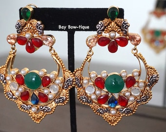 May include: A pair of large, ornate gold earrings with red, green, white, and blue gemstones. The earrings have a crescent moon shape and are decorated with intricate details.