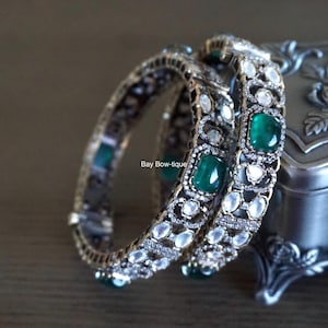 May include: Two silver bangles with intricate designs featuring clear and green gemstones. The bangles are resting on a silver box with floral engravings.