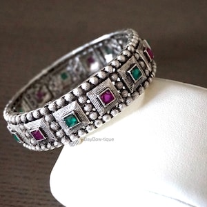 May include: Silver bangle bracelet with green and pink gemstones set in square bezels. The bracelet is decorated with small silver beads.