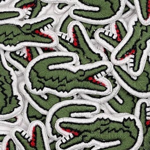 May include: A pile of green embroidered alligator patches with white outlines and red details in the mouth. The patches are arranged on a white surface, forming a repeating pattern. These patches are likely for clothing or accessories.