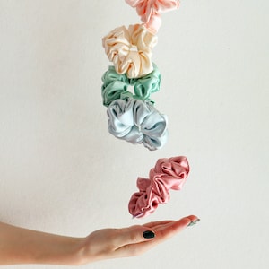 Könnte beinhalten: Eine Auswahl an Seiden-Haargummis in Pastellfarben wie Rosa, Creme, Mintgrün und Blau. Die Haargummis scheinen in der Luft zu schweben, eines davon liegt auf einer offenen Hand. Der Hintergrund ist weiß.
