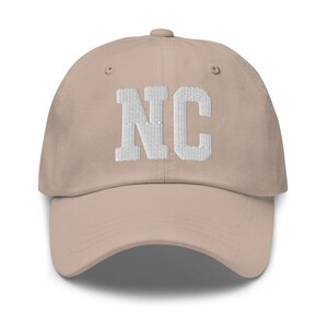 May include: A beige baseball cap featuring the embroidered white letters "NC". The cap has a curved peak and an adjustable strap. The letters are in a block style.