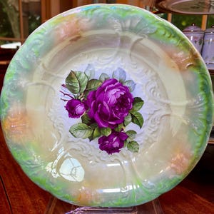May include: A vintage porcelain plate with a green iridescent glaze and a floral design featuring purple roses. The plate is on a clear acrylic stand.