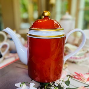 May include: A red and white ceramic teapot with gold accents. The teapot has a rounded body, a curved spout, and a handle. The lid is red with a gold knob. The teapot is sitting on a table with flowers.