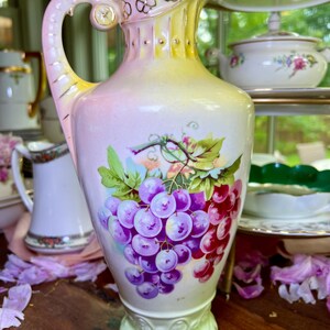 May include: A vintage porcelain pitcher with a delicate floral design and a cluster of grapes painted on the side. The pitcher has a light pink and yellow gradient and gold accents.