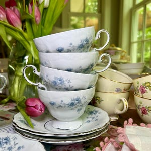 May include: A stack of white teacups and saucers with a blue floral design. The teacups have ornate handles and are stacked on top of each other. A bouquet of pink tulips is in the background.