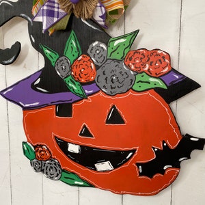 Op de afbeelding: Een houten Halloween-deurhanger in de vorm van een pompoen met een heksenhoed erop. De pompoen is oranje met een zwarte omtrek en een vriendelijk gezicht. De hoed is zwart met een paarse rand en groene bladeren rond de basis. Er zijn ook zwarte vleermuizen en bloemen op de hanger.