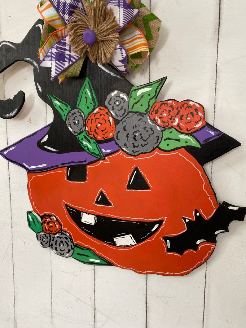 Op de afbeelding: Een houten Halloween-deurhanger in de vorm van een pompoen met een heksenhoed erop. De pompoen is oranje met een zwarte omtrek en een vriendelijk gezicht. De hoed is zwart met een paarse rand en groene bladeren rond de basis. Er zijn ook zwarte vleermuizen en bloemen op de hanger.