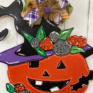 Op de afbeelding: Een houten Halloween-pompoen deurhanger met een zwarte heksenhoed, een paarse rand en een oranje gezicht. De pompoen is versierd met groene bladeren en zwarte en oranje bloemen. Een geruite en groene strik met een jute centrum is bovenaan bevestigd.
