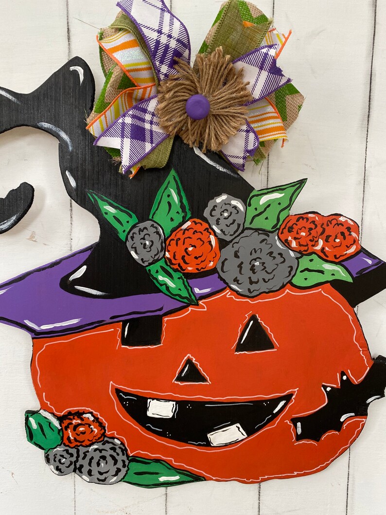 Op de afbeelding: Een houten Halloween-pompoen deurhanger met een zwarte heksenhoed, een paarse rand en een oranje gezicht. De pompoen is versierd met groene bladeren en zwarte en oranje bloemen. Een geruite en groene strik met een jute centrum is bovenaan bevestigd.