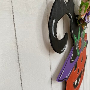 Op de afbeelding: Een houten heksendeurhanger met een zwarte hoed, een oranje en paarse pompoenkop en groene en witte bloemmotieven.