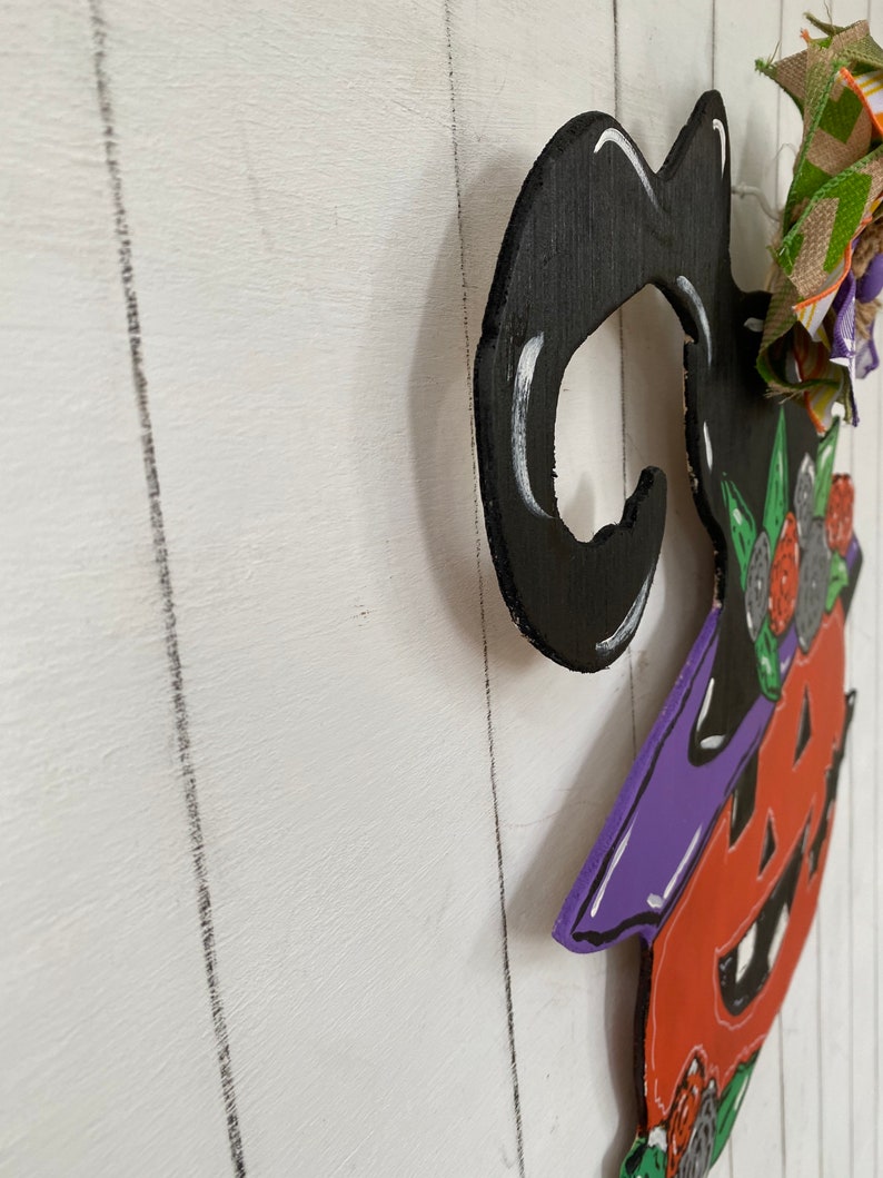 Op de afbeelding: Een houten heksendeurhanger met een zwarte hoed, een oranje en paarse pompoenkop en groene en witte bloemmotieven.