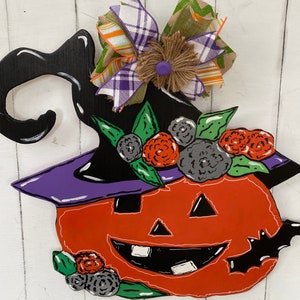 Op de afbeelding: Een houten Halloween-deurhanger in de vorm van een pompoen die een heksenhoed draagt met een paarse rand en een zwarte strik met groen-oranje ruitjes. De pompoen heeft een zwart-oranje gezicht met een brede glimlach en een zwarte vleermuisvleugel aan de zijkant. De hoed is versierd met groene bladeren en zwarte en rode bloemen.
