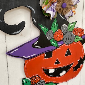 Op de afbeelding: Een houten Halloween-deurhanger in de vorm van een pompoen met een heksenhoed. De pompoen is oranje met een zwart gezicht en een paarse hoed. De hoed is versierd met groene bladeren en zwarte en rode bloemen.