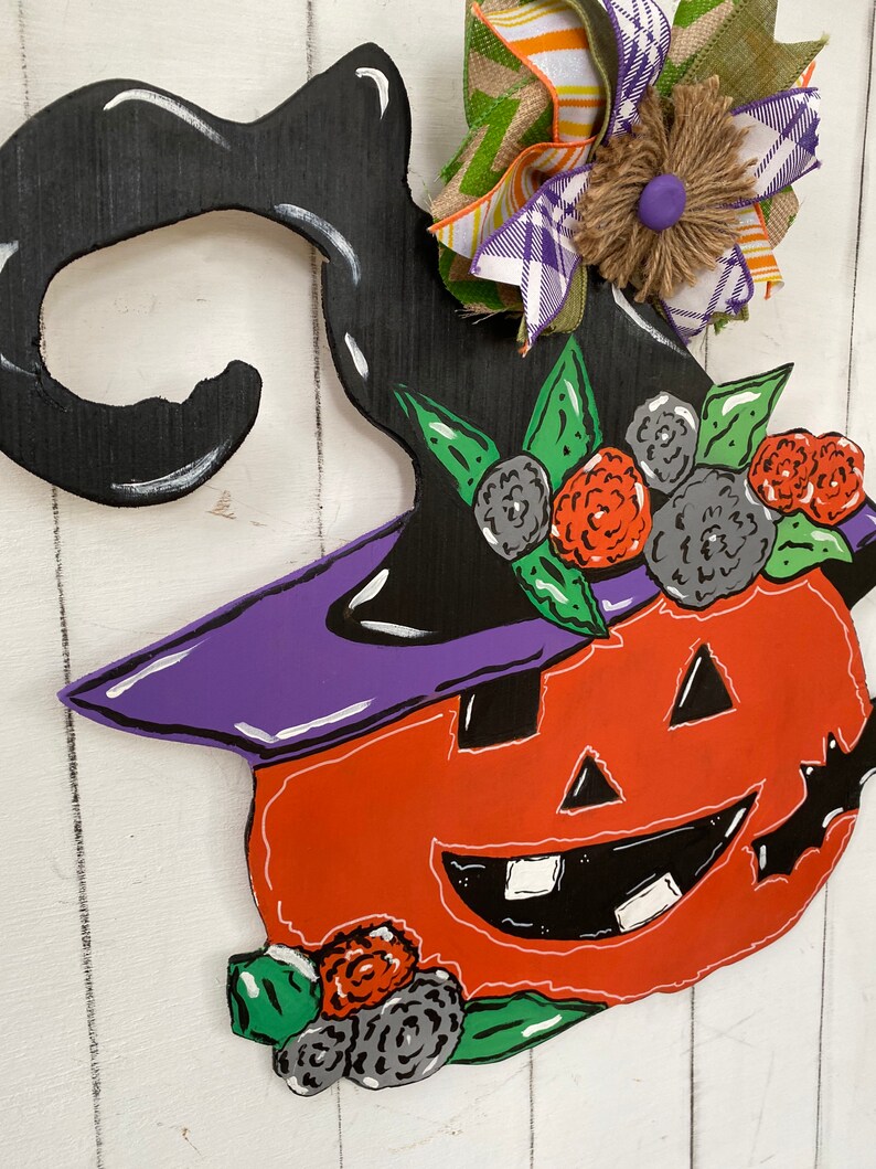 Op de afbeelding: Een houten Halloween-deurhanger in de vorm van een pompoen met een heksenhoed. De pompoen is oranje met een zwart gezicht en een paarse hoed. De hoed is versierd met groene bladeren en zwarte en rode bloemen.
