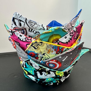 May include: A stack of five fabric bowl cozies with various colorful patterns. The patterns include comic book characters, avocados, and cartoon figures. Each cozy has a different color trim, including blue, pink, and yellow.