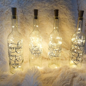 Könnte beinhalten: Vier durchsichtige Glasflaschen mit Korkverschlüssen und weißen Lichterketten im Inneren. Jede Flasche hat einen schwarzen Vinyl-Aufkleber mit einem anderen deutschen Spruch über die Familie. Die Flaschen stehen auf einer weißen, flauschigen Oberfläche.