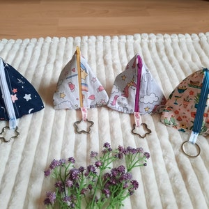 Könnte beinhalten: Vier dreieckige Reißverschlusstaschen mit verschiedenen bunten Mustern und sternförmigen Schlüsselanhängern. Die Taschen sind aus Stoff und haben einen Reißverschluss. Die Muster umfassen einen blauen und weißen Sternenhimmel, ein weißes und rosa Einhorn, einen weißen und gelben Regenbogen und einen rosa und grünen tropischen Druck.