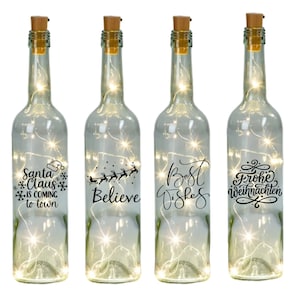 Könnte beinhalten: Vier durchsichtige Glasflaschen mit Korkverschlüssen und weißen Lichtern im Inneren. Jede Flasche hat ein anderes weihnachtliches Design: "Santa Claus is coming to town", "Believe", "Best Wishes" und "Frohe Weihnachten".
