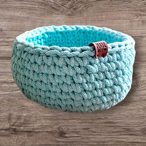 Könnte beinhalten: Ein hellblauer gehäkelter Korb mit einem braunen Lederanhänger, auf dem "Made with love" steht.