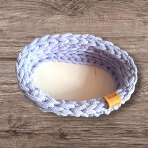 Könnte beinhalten: Ein helllila gestrickter Korb mit einem braunen Lederanhänger, auf dem "Little Hands" steht.