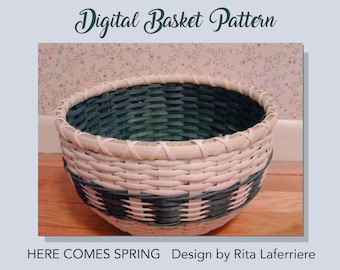 May include: A woven basket with a white and green pattern. The basket is round and has a wide base. The basket is labelled "Here Comes Spring" and "Design by Rita Laferriere".