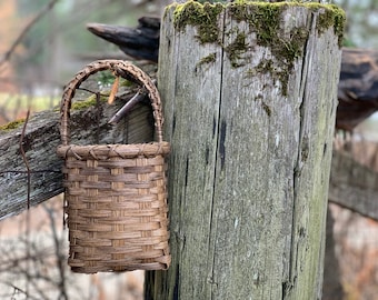 May include: A woven brown basket with a handle hangs on a weathered wooden fence post. The basket is made of natural materials and has a rustic appearance.