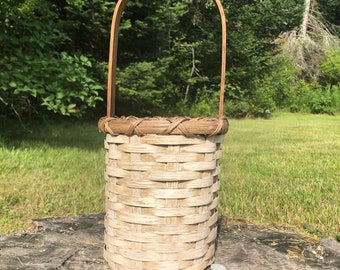 May include: A woven basket with a rounded handle. The basket is cylindrical with a light brown color and a woven pattern. The handle is made of a darker brown wood. The basket is sitting on a tree stump in front of a green background.