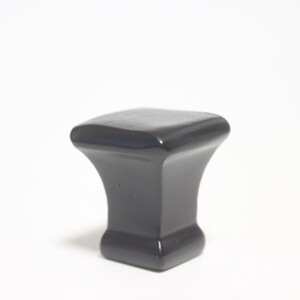 May include: A dark gray cabinet knob with a square top and a flared base. The knob has a smooth, glossy finish and is designed for furniture. The hardware is shown against a white background.