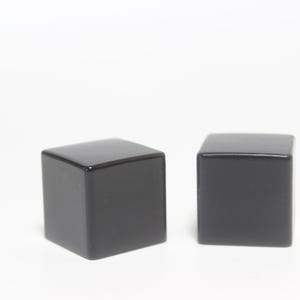May include: Two black, square-shaped objects with a glossy finish are displayed against a white background. The objects are identical cubes, each with a smooth, reflective surface. The lighting highlights their geometric form.