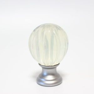 May include: A decorative curtain rod finial with a clear, ribbed, orb-shaped top. The orb is mounted on a silver-colored base. The finial is designed to be mounted on a curtain rod.