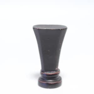 Puede incluir: Un sello de cera negro de estilo vintage con un cuerpo acampanado y facetado y una parte superior plana. La base tiene un diseño decorativo y redondeado. Los bordes muestran indicios de pintura roja, lo que sugiere edad y uso. El sello está sobre un fondo blanco.