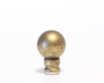 May include: A bronze-colored, spherical finial with a textured surface. The finial sits atop a cylindrical base with a tiered design. The metal finish has an aged appearance, suggesting a vintage or antique style. The finial is likely for a lamp or curtain rod.