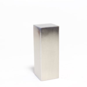 Puede incluir: Un objeto rectangular plateado metálico con una parte superior cuadrada y una superficie lisa y reflectante. El objeto está de pie sobre un fondo blanco, proyectando una sombra sutil.