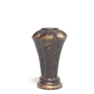 Puede incluir: Un pomo decorativo de color bronce con un diseño estriado. El pomo tiene una parte superior redondeada con un patrón floral y una apariencia envejecida y texturizada. La base tiene un diseño circular con un borde dorado.