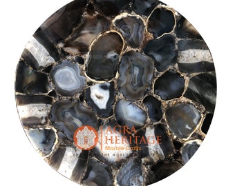 May include: A round, polished stone tabletop with a mosaic pattern of gray, black, and white agate slices. The agate pieces are outlined with a gold-colored material. The logo "AGRA HERITAGE Marble Crafts" is visible.