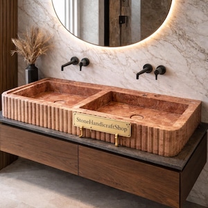 Peut inclure: Un double lavabo en pierre brun-rouge avec un extérieur nervuré. Le lavabo est monté sur un meuble en bois foncé. Au-dessus du lavabo se trouve un miroir rond avec un bord lumineux et des robinets noirs. Une enseigne indique "StoneHandicraftShop".