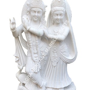 May include: A white marble sculpture depicting two figures, one playing a flute, the other standing beside them. The sculpture features intricate details, including ornate headdresses and flowing garments. A peacock is at the base. The text "AGRA HERITAGE MARBLE CRAFTS" is visible.
