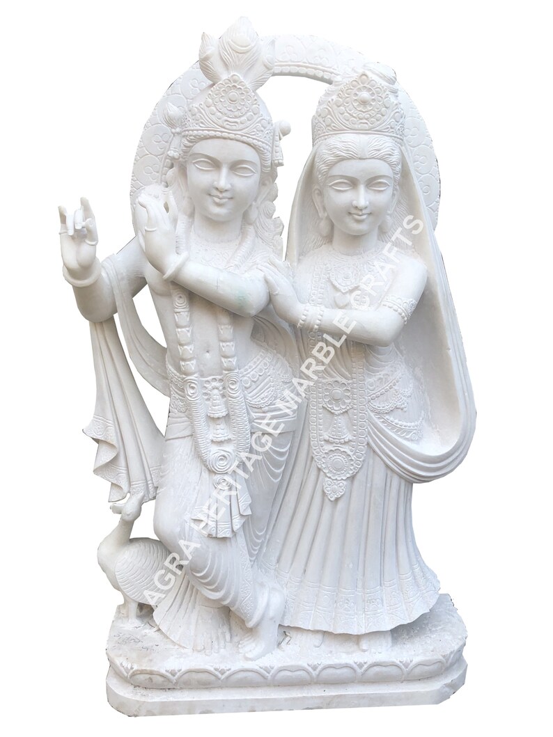 May include: A white marble sculpture depicting two figures, one playing a flute, the other standing beside them. The sculpture features intricate details, including ornate headdresses and flowing garments. A peacock is at the base. The text "AGRA HERITAGE MARBLE CRAFTS" is visible.