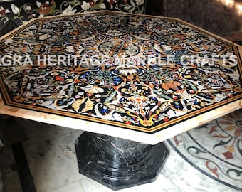 May include: Octagonal table with a black marble base and a colorful inlaid top featuring a floral design. The table top is made of various types of stone and has a beige border. The text "AGRA HERITAGE MARBLE CRAFTS" is visible on the table top.